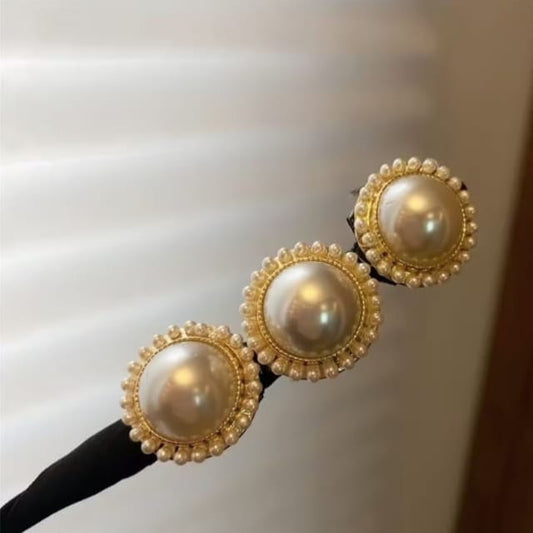 A close-up of a black hair accessory featuring three large round pearl-like centers surrounded by gold-tone beaded borders, shown against a softly blurred indoor background.