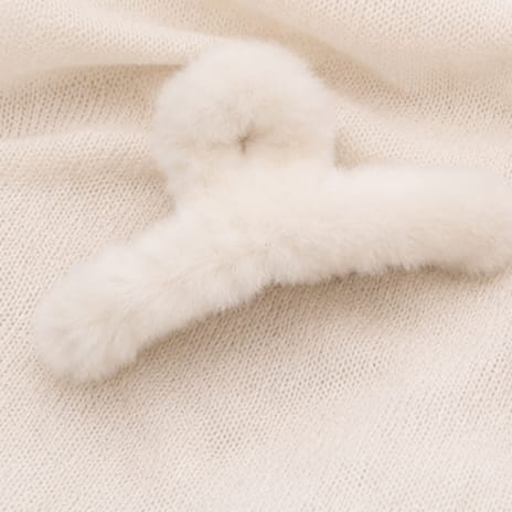 A fluffy cream faux fur hair claw clip resting on a textured cream knit fabric background.