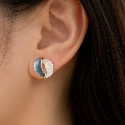 A close-up of a person's ear wearing a small crescent moon stud earring with gold, silver, and blue enamel details.