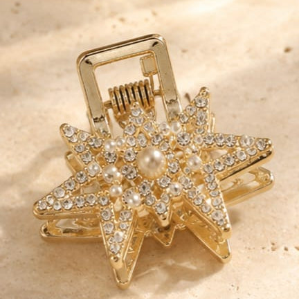 A gold-toned star-shaped hair claw clip embellished with clear rhinestones and a central pearl, resting on a textured beige stone surface.