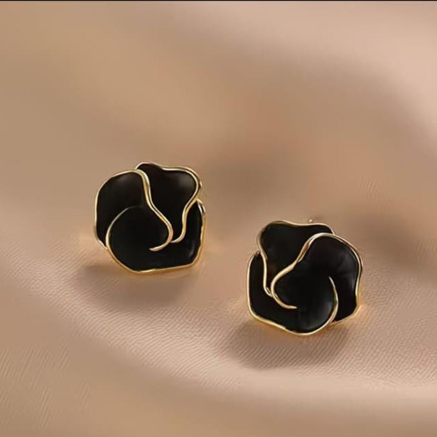A pair of black and gold rose-shaped stud earrings displayed on a soft beige fabric background.