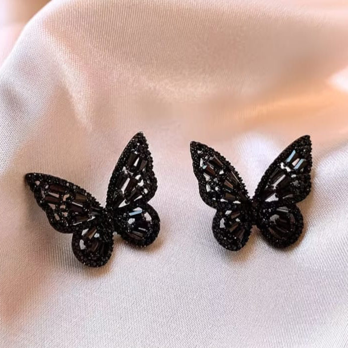 A pair of black butterfly-shaped stud earrings with sparkling clear rhinestone accents, displayed on a soft, draped cream satin fabric.