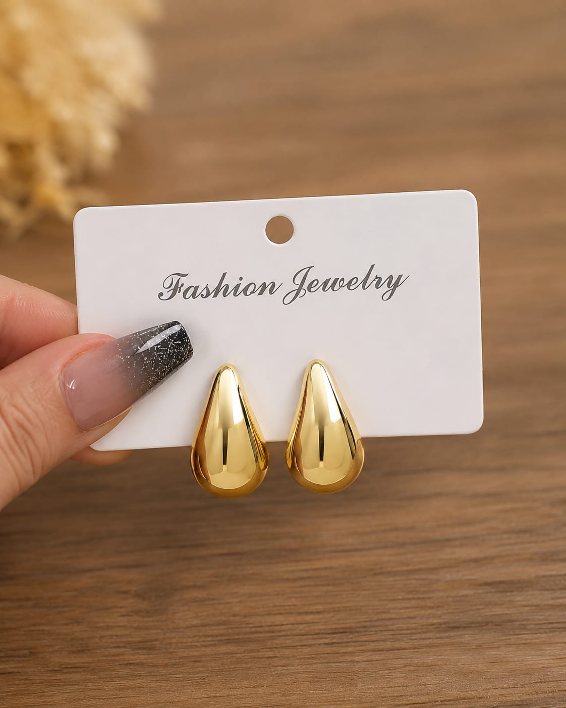 A pair of shiny gold teardrop-shaped stud earrings displayed on a white card labeled 'Fashion Jewelry,' held by a hand with a glittery black-and-nude manicure against a wooden surface.