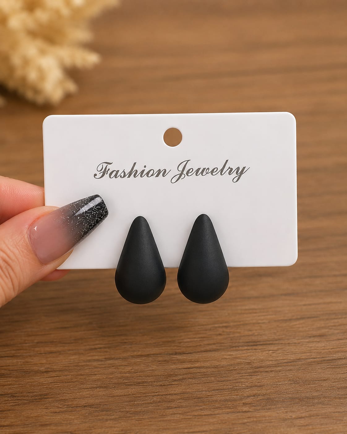 A pair of matte black teardrop-shaped stud earrings displayed on a white card labeled 'Fashion Jewelry,' held by a hand with a glittery ombre manicure against a wooden tabletop background.