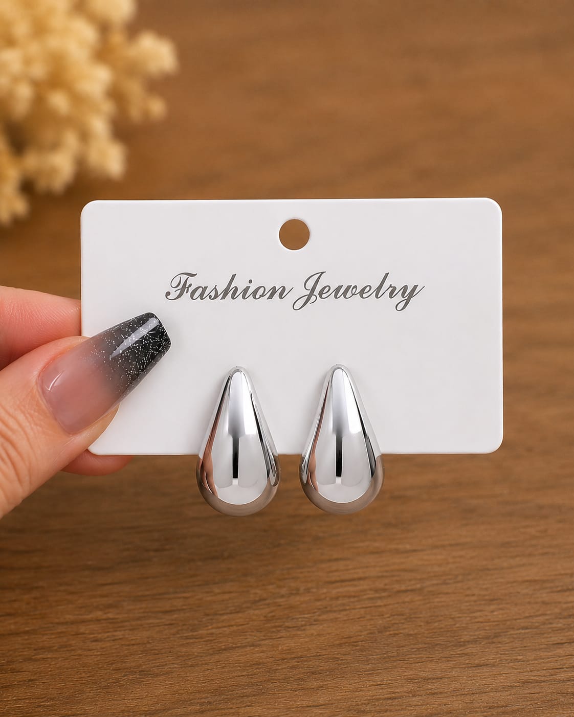 A pair of shiny silver teardrop-shaped stud earrings displayed on a white card labeled 'Fashion Jewelry,' held by a hand with glittery nail polish against a warm wooden background.