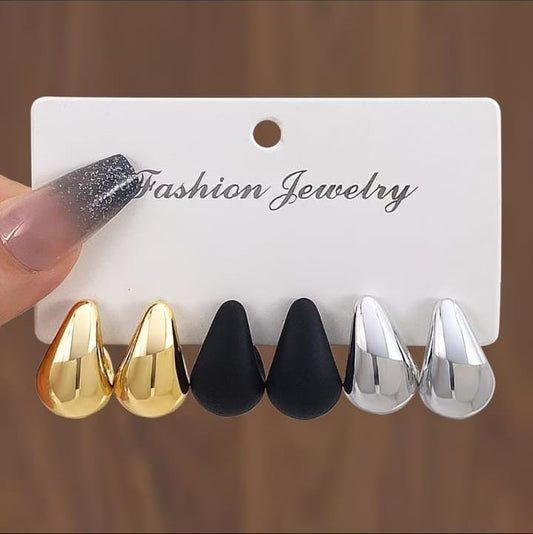 Six teardrop-shaped fashion jewelry studs in gold, matte black, and polished silver, displayed on a white card labeled 'Fashion Jewelry' with a hand holding the left side.