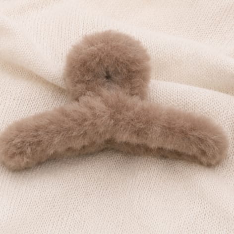 A fluffy faux-fur hair claw clip in a light taupe-brown color, displayed on a cream-colored knit fabric background.