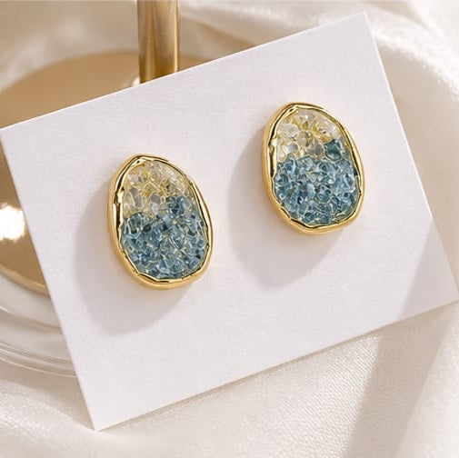 A pair of gold-framed stud earrings with textured blue and cream resin centers, displayed on a small white card over a soft white fabric background.