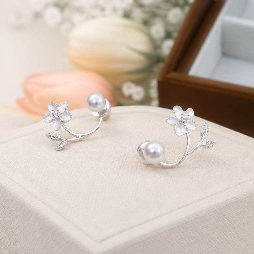 A pair of silver-toned ear cuffs styled as delicate flowers with white pearls and small clear crystals, displayed on a cream jewelry box.