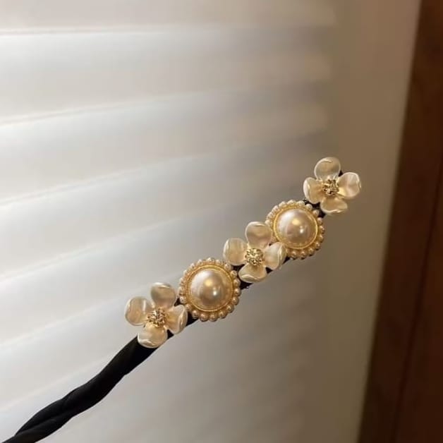 A black hair accessory headband decorated with three gold-tone floral charms, each featuring a faux pearl center, resting against a soft-focus background of white window blinds.
