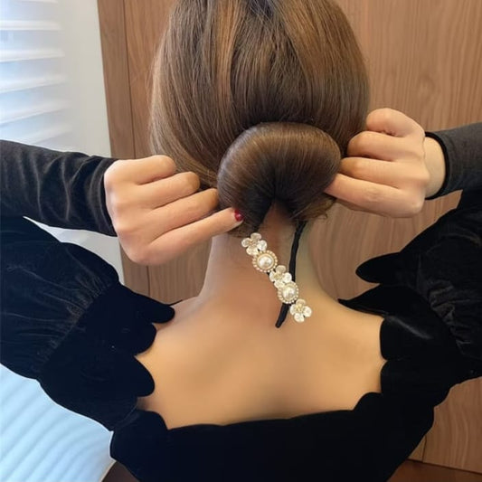 A woman shown from behind with her hair styled in a low bun, wearing a decorative hair accessory with clear rhinestones and pearls. She is wearing a black velvet top with ruffled sleeves.