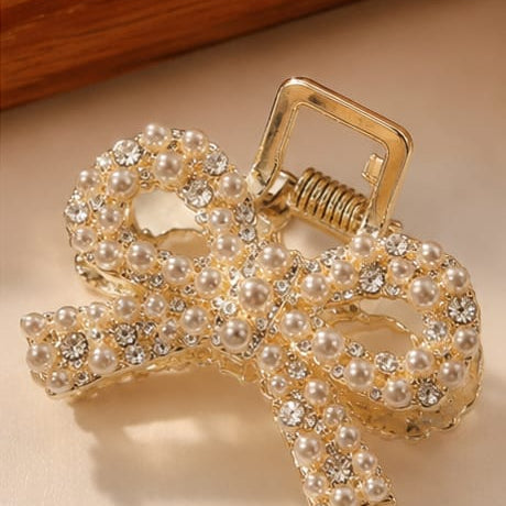 A gold bow-shaped hair claw clip embellished with clear rhinestones, resting on a light beige surface with decorative fabric and wooden elements in the background.