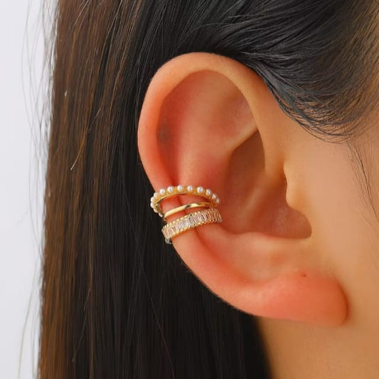 A close-up of a person's ear wearing a stack of three gold ear cuffs: one with small white pearls, one plain gold, and one set with clear rhinestones.