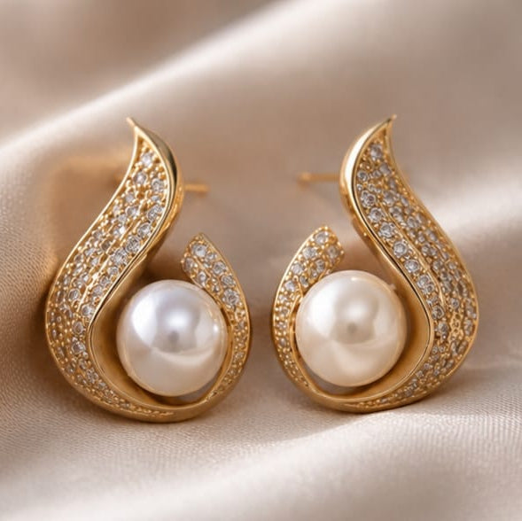A pair of gold teardrop-shaped earrings with white freshwater pearls and pavé-set clear stones, displayed on a soft beige satin fabric background.