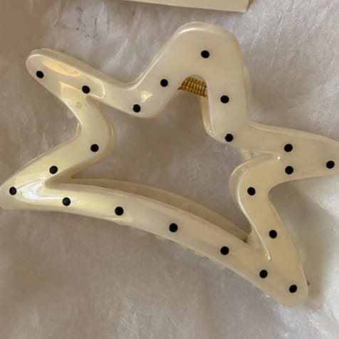 An off-white star-shaped hair claw clip decorated with small black polka dots, resting on a textured white fabric background.