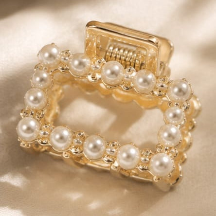 A gold square hair claw clip adorned with white faux pearls, resting on a beige satin fabric with soft shadows.