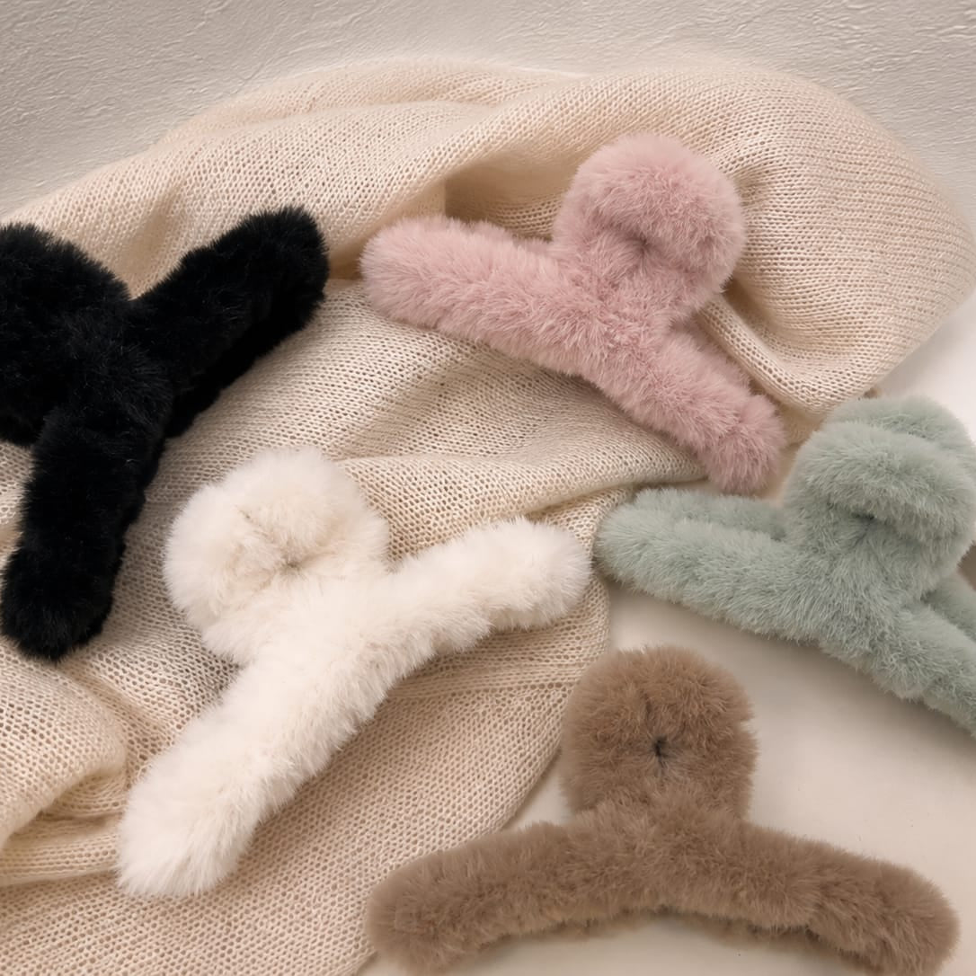 Five fluffy faux-fur claw hair clips in black, pink, white, sage green, and taupe, arranged on a cream-colored knit blanket.