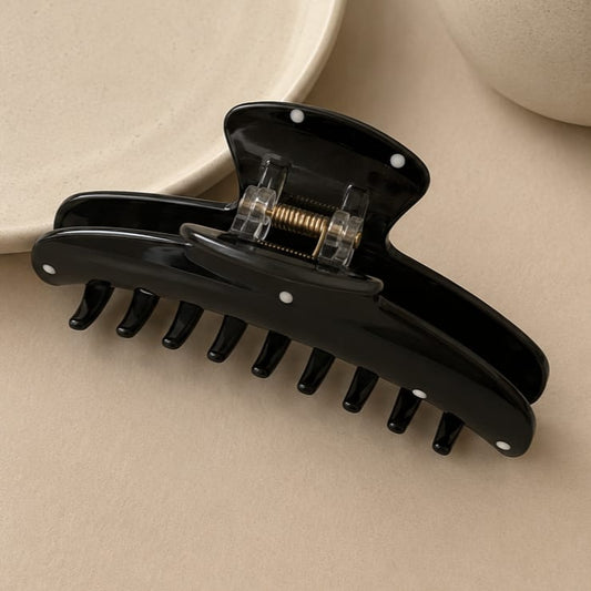A glossy black hair claw clip with clear plastic accents and small white dots, photographed on a warm beige surface with a softly blurred neutral background.