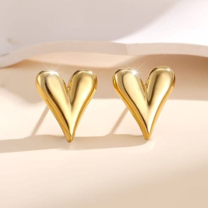 A pair of shiny gold heart-shaped stud earrings displayed on a warm beige surface with soft shadows.