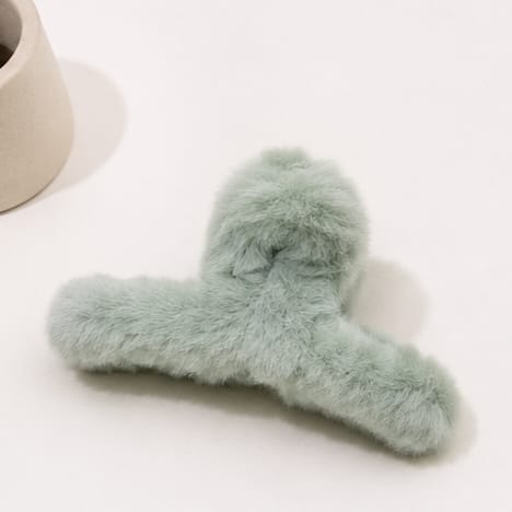A fluffy faux fur hair claw clip in a pale sage green color, displayed on a clean white surface with a neutral beige ceramic pot partially visible in the top left corner.