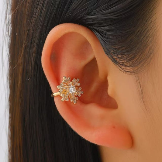 A close-up of a person's ear wearing a gold ear cuff earring adorned with small clear crystals in a floral or snowflake-like design.