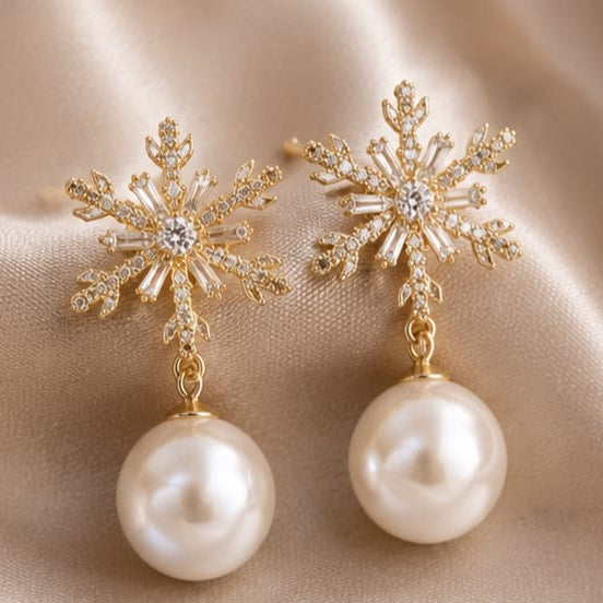A pair of gold snowflake-shaped drop earrings with clear rhinestones and a large white pearl dangling below, displayed on a beige satin fabric background.