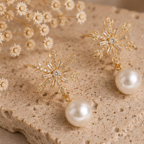 A pair of gold snowflake-shaped drop earrings with clear rhinestones and a single white pearl dangling below, displayed on a textured beige stone surface with dried white flowers in the background.