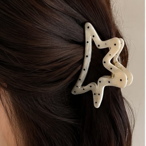 A white star-shaped hair claw clip with black polka dots, shown holding back dark brown hair on a model's head.