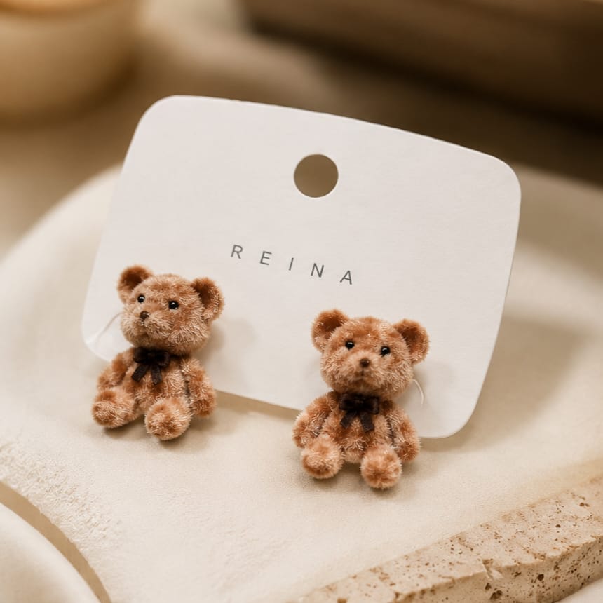 A pair of small teddy bear stud earrings on a white display card with the brand name REINA, photographed on a soft beige surface.