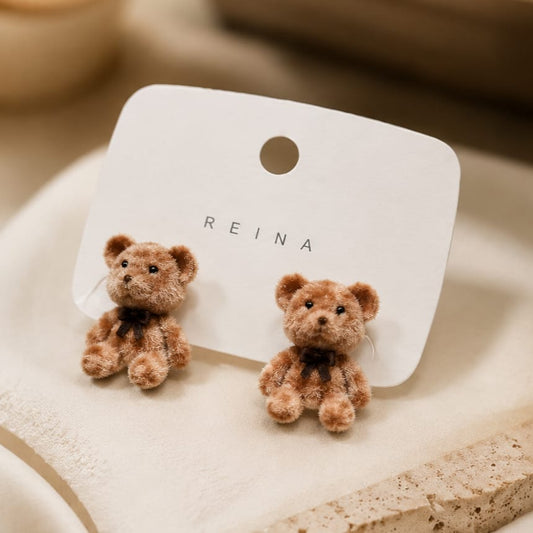A pair of small teddy bear stud earrings on a white display card with the brand name REINA, photographed on a soft beige surface.