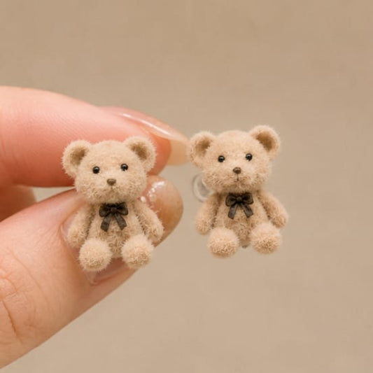 A close-up of a hand holding two small teddy bear stud earrings. The bears are light beige with black embroidered eyes, noses, and small black bows at their necks.