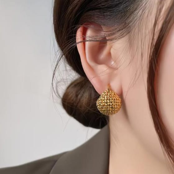 A close-up of a person's ear wearing a textured gold teardrop-shaped stud earring, with dark hair and a dark blazer visible against a light neutral background.