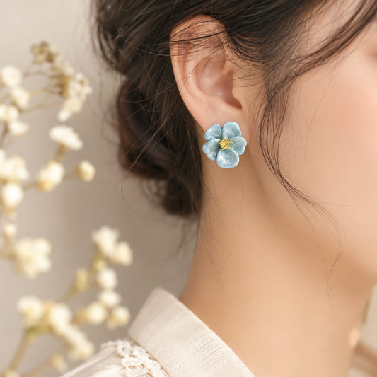 A close-up of a woman's ear wearing a light blue flower-shaped stud earring with a small gold center, against a soft beige background with blurred white flowers.