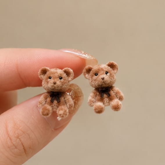 Two miniature brown teddy bear stud earrings held between fingers against a soft beige background.